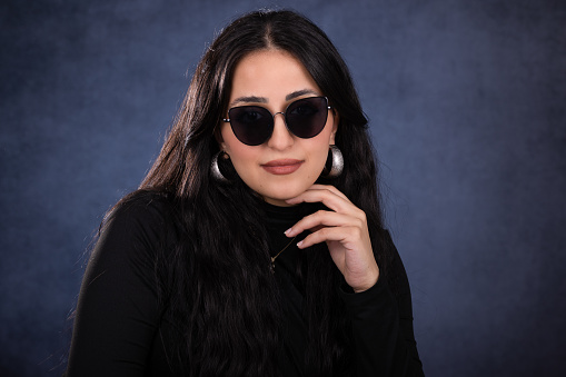 Young lady studio portrait over blue background. She looks serious while facing the camera while putting her hand on her chin. She wears a black sweater and black sunglasses.