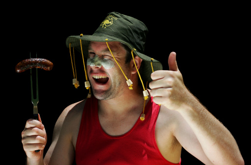 An Aussie man in a cork hat winks at the camera holding up a sausage on a fork in one hand and giving the thumbs up gesture with the other