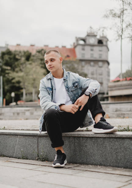 city solitude. thoughtful young man in a denim jacket. stylish brooding. young guy ponders life in his denim jacket. cityscape contemplation. young man in denim jacket lost in thought - bolero jacket imagens e fotografias de stock