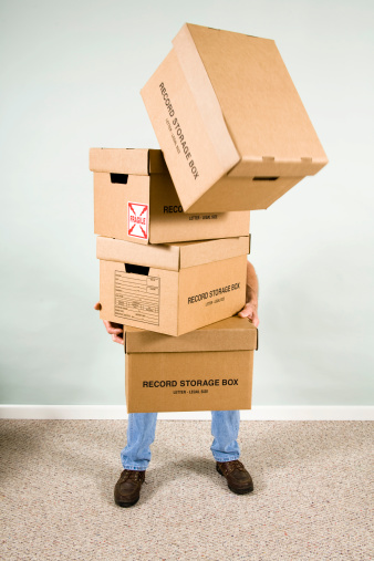 Man trying to carry too many cardboard boxes, top one is falling from stack.  Man holding boxes falling from the stack.  Indoors. 