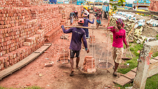 People offloading bricks from the boat, the image captured on May 29, 2022, from Amen Bazar, Bangladesh, where workers are busy unloading heavy bricks from a boat onto the dock. They carefully carry the bricks, one by one, from the boat's cargo hold to the dock, using a combination of teamwork and manual labor. The air is filled with the sounds of clinking bricks and the occasional shouts of coordination as they efficiently transfer the construction materials to the awaiting area on the shore. This scene depicts the physical effort and cooperation required in unloading bricks from a boat, a common sight at construction sites and ports around the world.