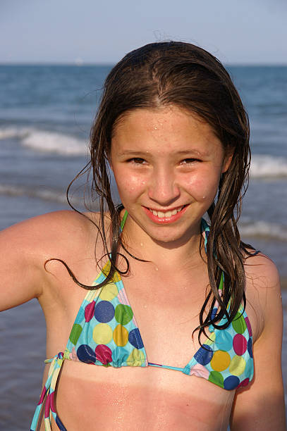 A young Asian teenage girl in a white dress smiles and looks at the camera while standing on the beach.