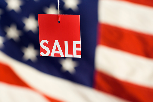 A hanging sale tag against a USA red, white, and blue American flag background in soft focus. Price tag label for bargain shopping on the Fourth of July, Memorial Day, or other American holidays and events, or for marketing discounted merchandise to appease consumerism and promote retail business commercial activity.