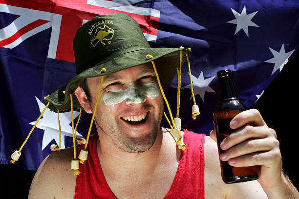 An Aussie man in a cork hat smiles at the camera holding up a beer
