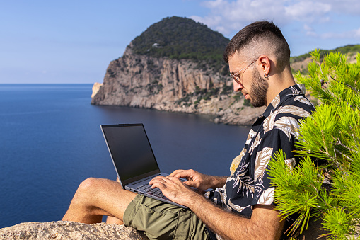 Freelancer working on a cliff with his laptop with the sea in the background. Digital nomad