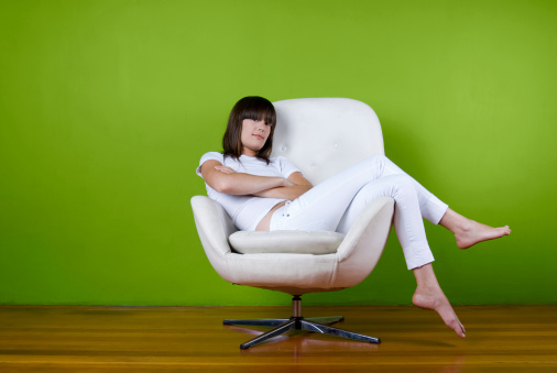 A young woman sits in a white mid century chair while dressed in white against a green background.