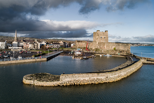 Carrickfergus, Northern Ireland, UK - February 1, 2020: Medieval Norman Castle, harbor with boat ramp and wave breaker in Carrickfergus near Belfast, Northern Ireland, UK. Aerial view in sunset light in winter. Town and stormy clouds in the background