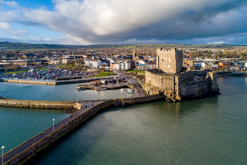 Carrickfergus, Northern Ireland, UK - February 1, 2020: Medieval Norman Castle, harbor with boat ramp and wave breaker in Carrickfergus near Belfast, Northern Ireland, UK. Aerial view in sunset light in winter. Town and stormy clouds in the background