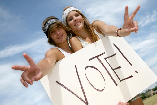Young Adult Hippie Couple Holding Vote Sign Making Peace Hand Gesture With Blue Sky As The Background.