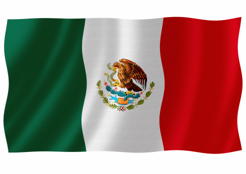 A large Mexican flag (Spanish: Bandera de México), lit with golden pre-sunset light, on a flag pole, with a blue sky, in the Zocalo, Mexico City