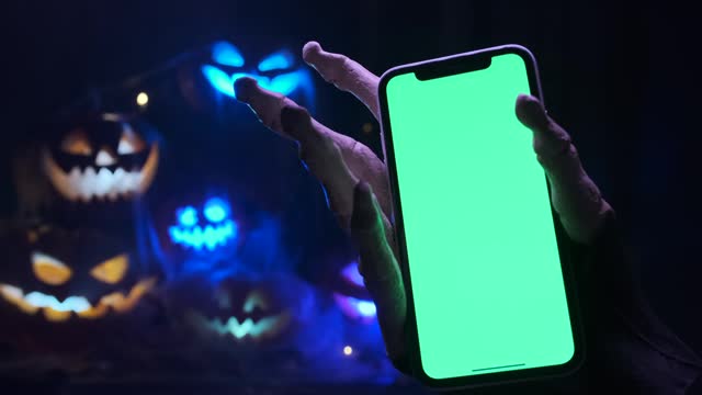 Spooky hand holding phone with chromakey on Halloween background of many lanterns made of glowing pumpkins