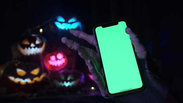 Scary skeletal monster hand holding smartphone with chromakey green screen against background of pyramid of Jack-o'-lanterns