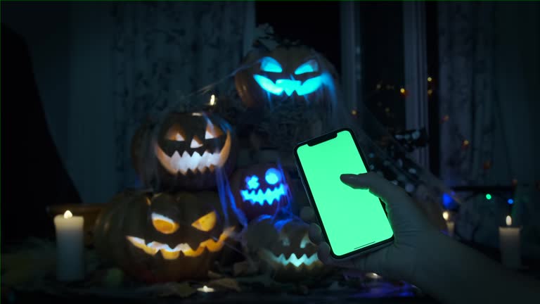 Hand holds phone with green screen touch on with finger, against background of pyramid of carved pumpkin Jack-o'-lanterns