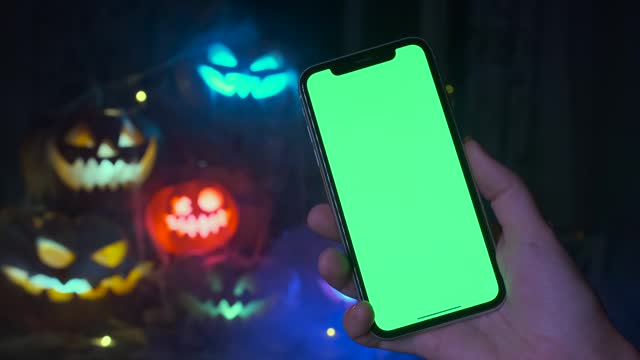 Hand holding phone with chromakey closeup against blurred festive background with bunch of gcarved pumpkins