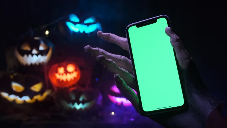 Creepy skeleton hand holding a phone with green screen at a party during Halloween closeup against blurred background