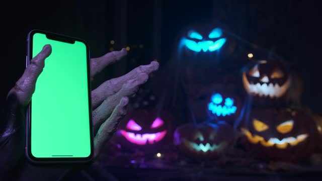 Close-up of skeletal fingers holding phone with chromakey calling to come to the party on Halloween holiday