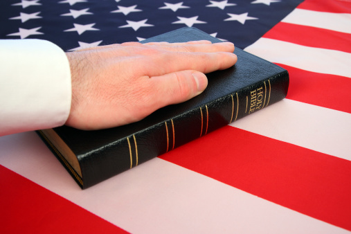 This color photo shows a hand resting on a black and gold bible as if taking an oath. The bible rests on an American flag background, giving a patriotic feel with stars, stripes, and red, white, and blue colors. Concept can symbolize presidential inauguration, court testimony, or other aspects of the American legal system.