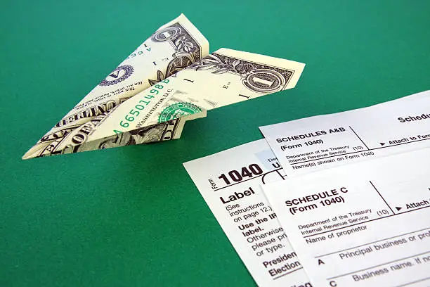 This color photo shows a sleek paper airplane made from a one-dollar bill ($1) over a dark green background. Parts of 1040 tax forms from the IRS, including Schedule C and Schedule A&B, are visible in the lower right corner of the image next to the paper airplane.