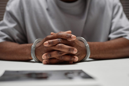 Close up of anonymous black man wearing handcuffs sitting at table in police department with hands clasped, copy space