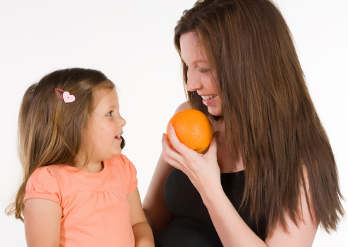 Mother and young daughter smile at each other while mom holds an orange