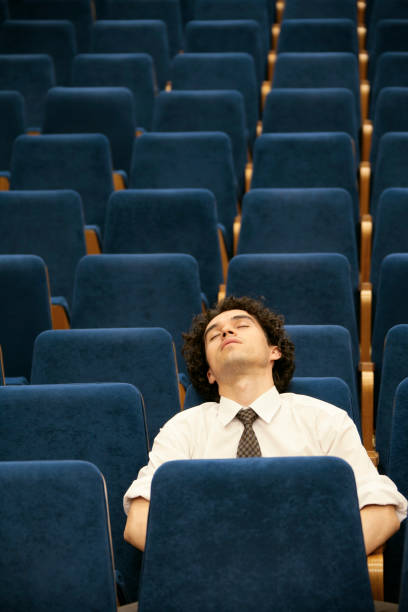 A businessman sleeping in a chair Young businessman sleeping in an empty conference hall (or movie/theatre) male college student sleeping through lecture stock pictures, royalty-free photos & images