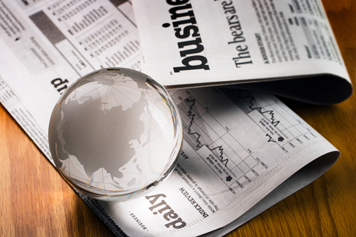 A glass globe paperweight resting on newspaper financial pages, representing international business markets and global finance. The earth sphere implies fortune telling economic trends and forecasting stock market activity for investment success in a recession or unstable market. China, India, and Japan are visible, for concepts of Asian world economy.