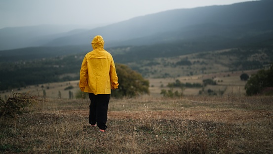 A woman is wearing a yellow raincoat and walking in nature and mountain during cold and rainy weather.