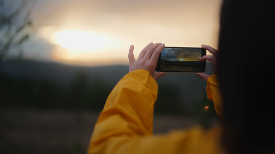 A woman in a yellow raincoat is appreciating the beauty of nature and taking photos with her mobile smart phone in nature in mountains in cold and rainy weather.