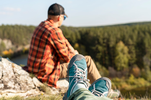 Women's legs in hiking boots relaxing on mountain near river in autumn and man in red plaid shirt looking at view Tourism