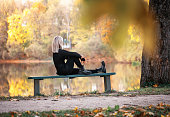 A blonde woman is relaxing on a sunny evening, sitting on a park bench near a pond and admiring the autumn view