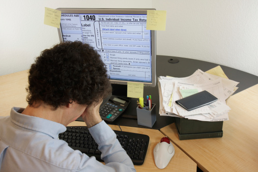 A distraught man contemplating doing his taxes on his computer with a shoebox of receipts in the background. IRS form 1040 and schedule A & B appear on the screen.