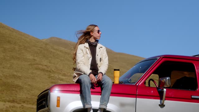 Attractive long-haired girl drinking a drink from a thermos of thermo mugs while standing near a vintage car against the backdrop of a mountain landscape.