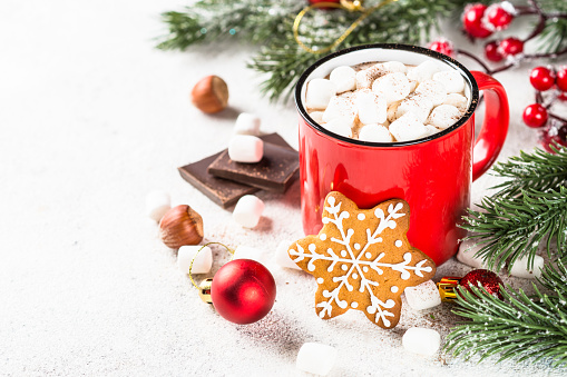 Hot chocolate with marshmallow and gingerbread cookies and holiday decorations at white table.