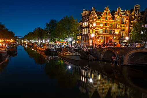 Netherlands. Summer night on the Amsterdam canal. Dancing Dutch houses with lights. Bar with outdoor tables. Moored houseboats and boats