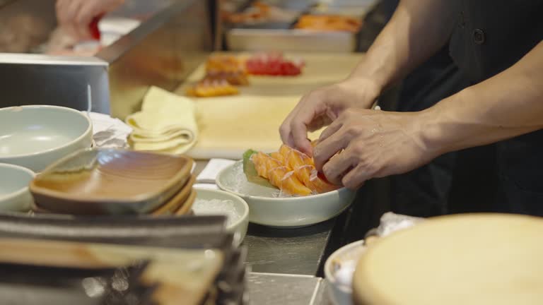 Chef's hands plating sliced salmon sashimi on ice in a decorated dish  while another chef filleting fish in a Japanese restaurant.