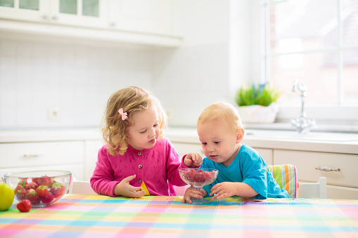 Child having breakfast. Kid drinking milk and eating cereal with fruit. Little boy and girl at white dining table in kitchen at window. Kids eat on sunny morning. Healthy balanced nutrition for kids.