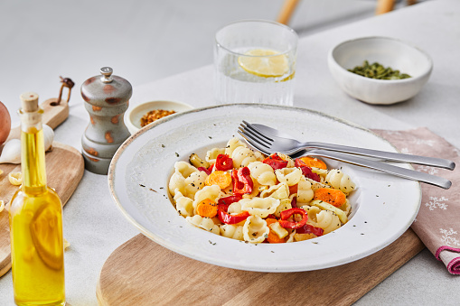 Healthy vegetarian pasta, made of zucchini, red pepper, carrot and garlic, served on an elegant ceramic plate, with fresh vegetables and herbs around, olive oil and pepper, on a white granite ceramic natural kitchen or restaurant table, representing a wellbeing and a healthy lifestyle, food indulgence, an image with a copy space