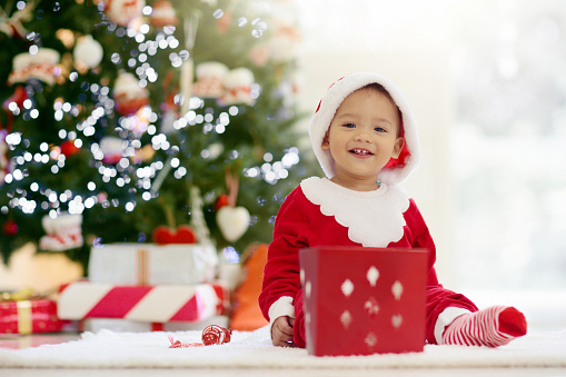 Child decorating Christmas tree at home. Little baby in pajamas with Xmas ornament. Asian family with kids celebrate winter holidays. Kids decorate living room and fireplace.