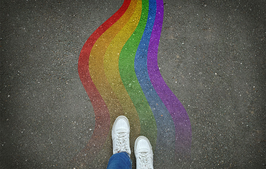 Legs in white sneakers in front of a wavy rainbow path on gray asphalt, top view. LGBT rainbow flag, gender identity concept