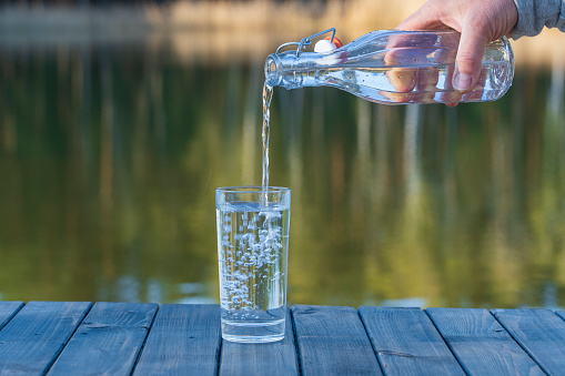 Pouring drink water into a glass from a bottle in the morning next to the lake and forest in spring time, close up. Nature and travel concept