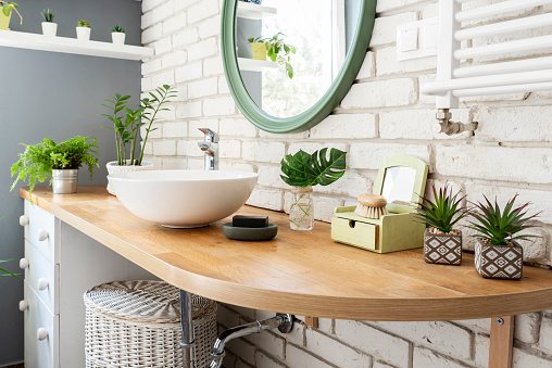 Interior of bathroom with a lot of plants on wooden counter and round mirror on white brick wall. Scandinavian style at home apartment.