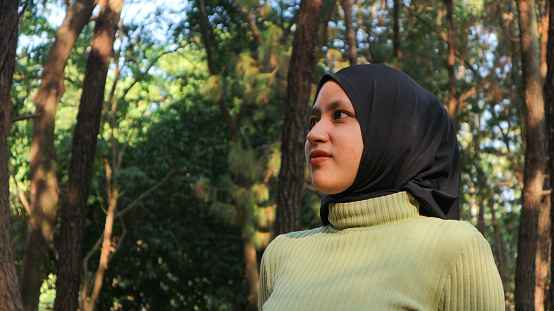 Close-up Muslim woman wearing hijab head scarf in pine forest. Modern muslim woman portrait, wearing black hijab in summer sunny park, looking at camera. Confident islamic girl.