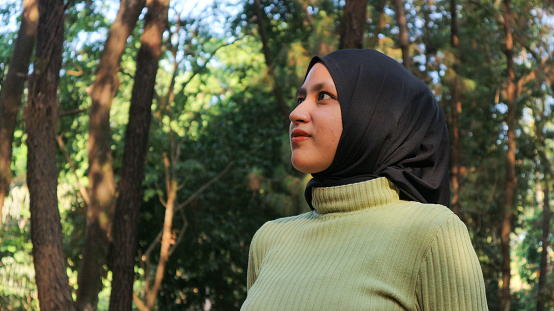 Close-up Muslim woman wearing hijab head scarf in pine forest. Modern muslim woman portrait, wearing black hijab in summer sunny park, looking side view. Confident islamic girl.
