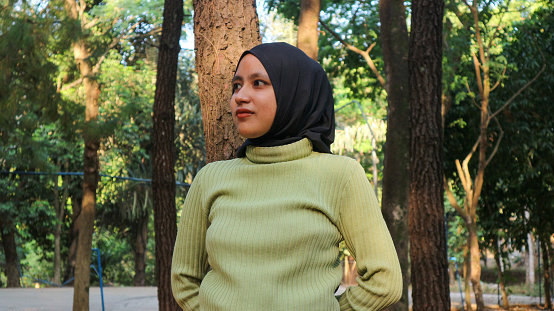 Close-up Muslim woman wearing hijab head scarf in pine forest. Modern muslim woman portrait, wearing black hijab in summer sunny park, looking side view. Confident islamic girl.