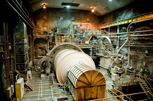 A twenty-foot diameter ball mill rotates at a copper mine's underground processing facility in Chile.  This process breaks copper-bearing ore from golf-ball sized pieces into dust. Dozens of other pumps, motors, ladders, cranes, lights, and scaffolding is visible surrounding this equipment.