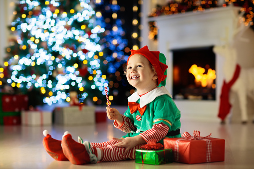 Child opening present at Christmas tree at home. Kid in elf costume with Xmas gifts and toys. Little baby boy with gift box and candy at fireplace. Family celebrating winter holidays. Home decoration.