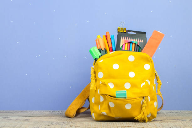 istock Yellow backpack with white polka dots with different colorful stationery on table. Purple background. Back to school. 1728100165