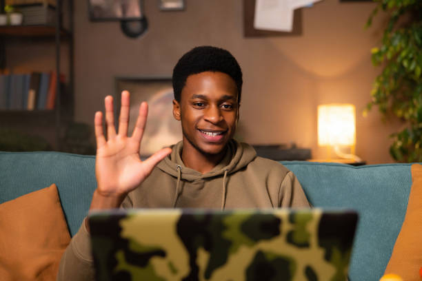 istock Happy dark skinned man student guy browsing the internet or social media, waving at someone video chatting with friends or family who live far away, waving at them as they greet him on the screen 1728165485