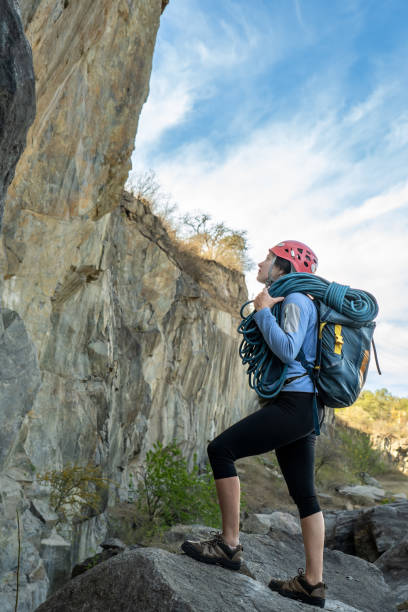 Female climber standing in front of a rock, carrying climbing rope, looking up, observing rocky mountain.