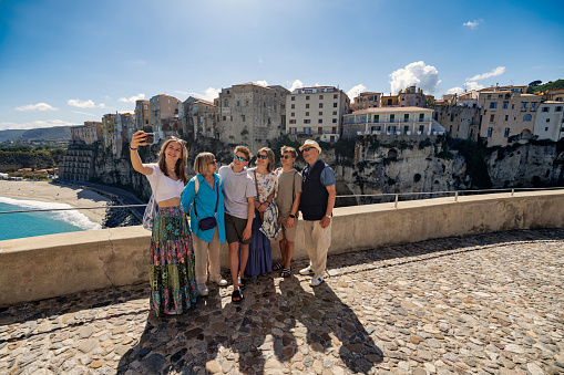 Multi generation family enjoying Calabrian town of Tropea on a sunny day. They are standing on the viewpoint by the Santuario di Santa Maria dell'Isola di Tropea and taking selfies. The beautiful cliff of Tropea is visible in the background.\nShot with Canon R5